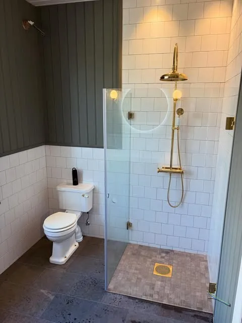 Färdigt badrum med vitt kakel, grön panel, dusch i mässing och glasvägg